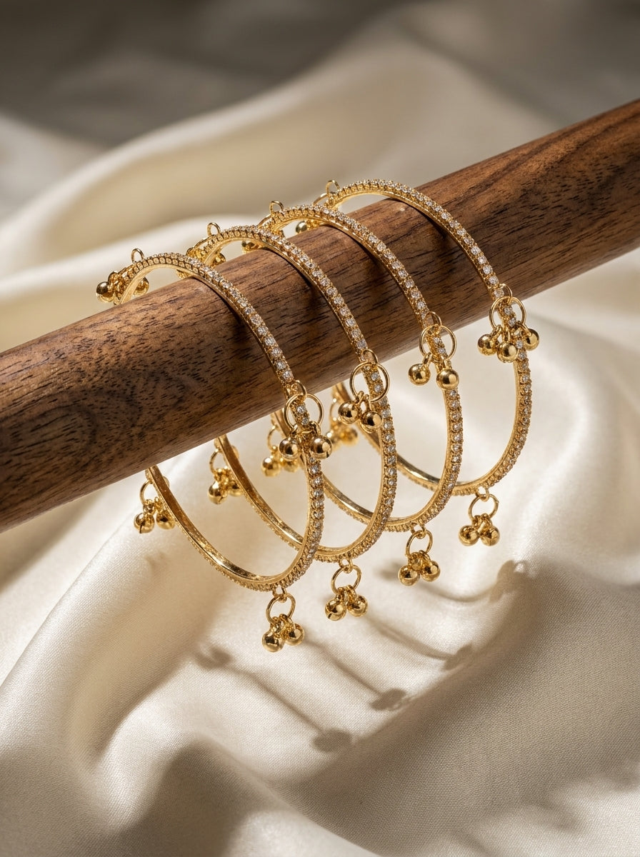 Set of four gold-plated CZ stone bangles with dangling ghungroo bell charms displayed on a wooden rod — Behuli Australia