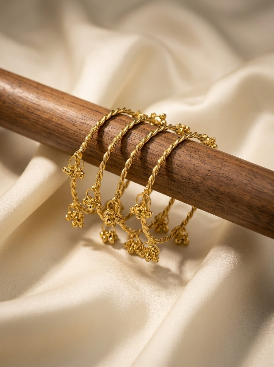 Set of four gold-tone twisted rope bangles with ornate floral ghungroo bell charms displayed on a wooden rod — Behuli
