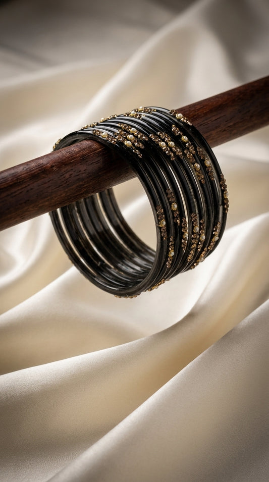 Stacked black bangles with gold stone and pearl detailing draped on a wooden rod against ivory fabric