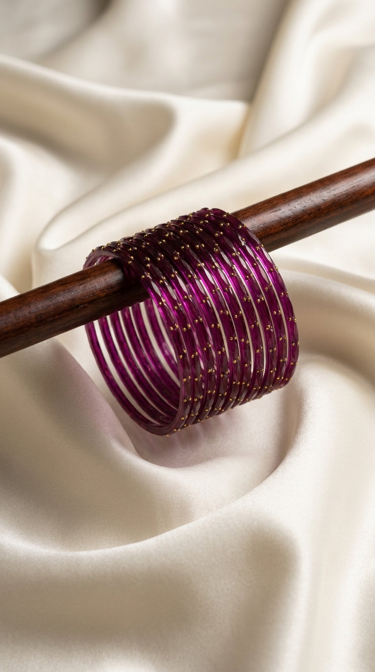 Set of 12 fuchsia glass bangles with gold-tone dot detailing displayed on a wooden rod against ivory silk — Behuli Australia