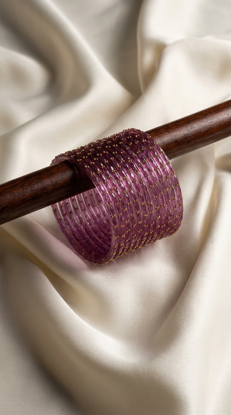 Stack of 12 purple bangles with gold-tone bead detailing displayed on a wooden rod against ivory silk — Behuli Australia