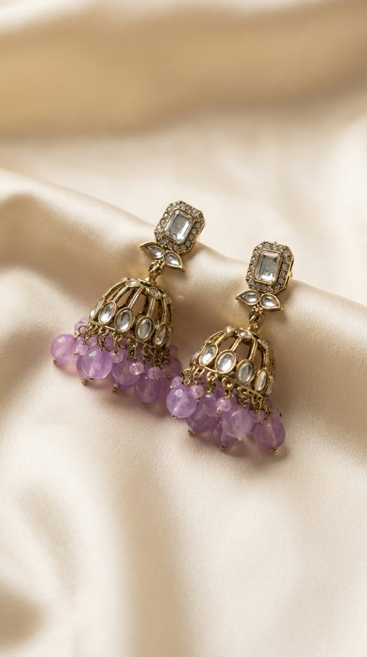 Gold-toned kundan jhumka earrings with cage dome, oval stone setting and cascading lavender bead drops on ivory silk —