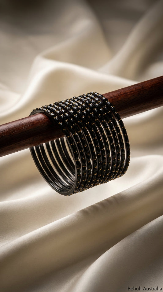 Set of 12 black bangles with gold bead detailing stacked on a wooden rod against ivory satin fabric