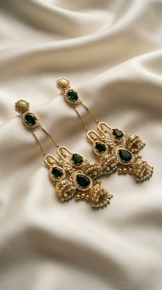 Gold-tone layered jhumka drop earrings with emerald green teardrop stones and ivory pearl bead clusters on cream silk —